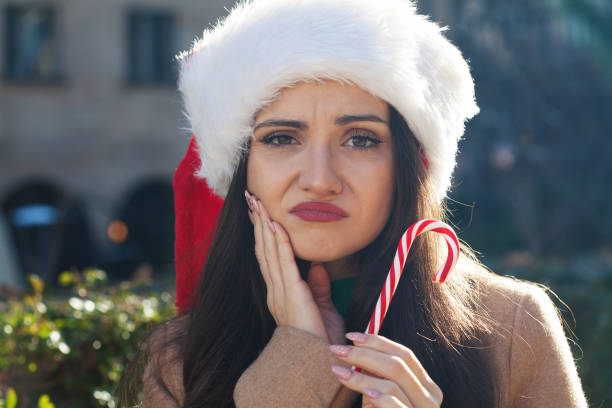 a woman wearing a Santa hat, touching her jaw due to toothache while holding a candy cane